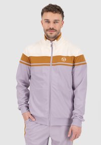 Trackjakke i lavendel og creme, med en brun stribe, fuld lynlås, høj krave og logo på brystet. Blødt stof med struktureret finish.
