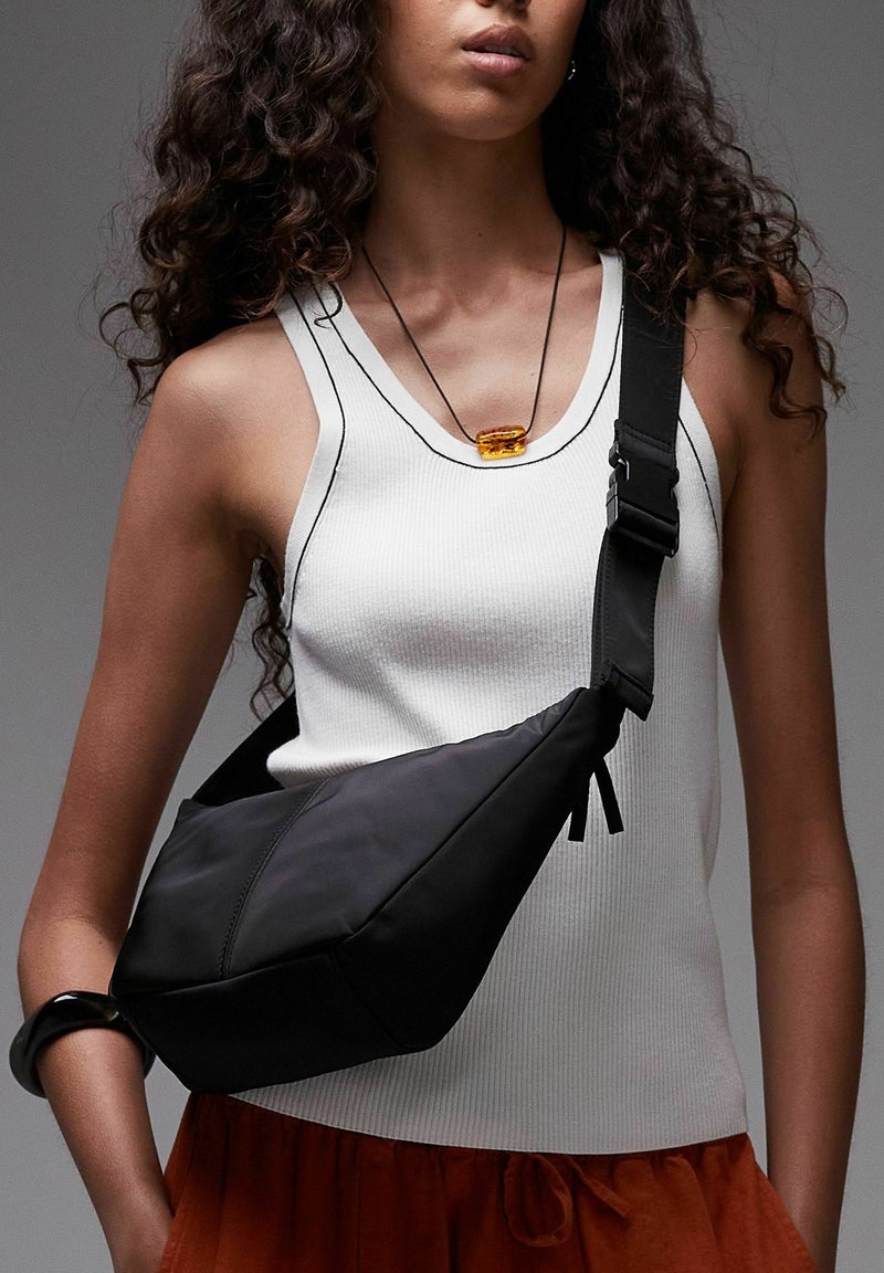 Vrouw draagend een witte mouwloze top, zwarte schoudertas, amberkleurige hangers ketting en een zwarte armband om haar linkerpols.