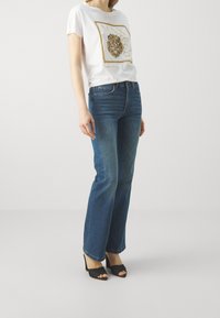 Camiseta blanca de manga corta con un gráfico de león dorado, combinada con unos vaqueros de corte relajado azules y unas sandalias de tacón alto negras.
