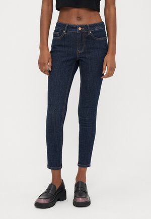 Vaqueros ajustados azul oscuro usados por una persona de pie, combinados con mocasines negros gruesos y una camiseta corta negra.