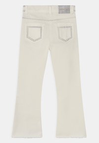 Jasno kremowe dżinsy w fasonie dzwonów z srebrnymi ćwiekami na kieszeniach i pasku, z surowym wykończeniem dołu dla nowoczesnego wyglądu.