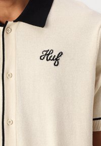 Cremefarbener Cardigan mit einem schwarzen gerippten Kragen und einem kleinen gestickten "Huf"-Logo. Verfügt über Knöpfe auf der Vorderseite und gerippte Bündchen.
