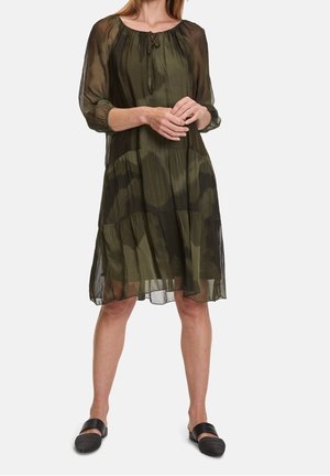 Femme portant une robe fluide vert olive transparente avec des volants froncés et manches 3/4, associée à des chaussures tissées noires sans lacets.