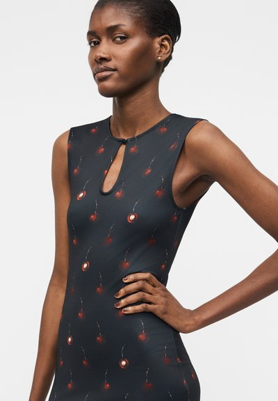 Kobieta w czarnej sukience bez rękawów z małymi czerwonymi wzorami wiśni i dekoltem w kształcie łezki, stojąca z jedną dłonią na biodrze.
