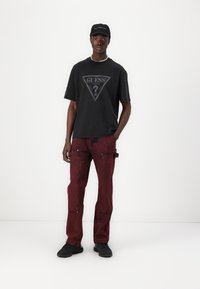Sort bomulds t-shirt med trekantet gråt logo, parret med røde slidte denimbukser. Sorte sneakers og en matchende kasket fuldender outfittet.