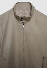 Veste beige avec col montant, fermeture éclair avant et deux poches latérales. Présente une texture lisse et mate ainsi qu'un design minimaliste.