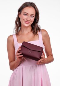 Bordó crossbody taška s texturovaným povrchem, magnetickým uzávěrem a logem; držena osobou ve světlém růžovém šatu.