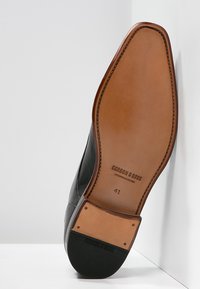 Chaussure en cuir noir avec un dessus lisse, un bout rond et une semelle en cuir brun. Marquage notable sur la semelle et indicateur de taille 41.