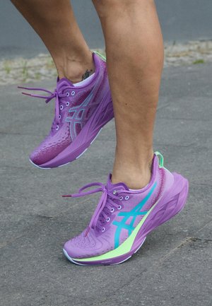 Personne portant des chaussures de course violettes et bleu sarcelle marchant sur un trottoir gris avec un pied légèrement levé.