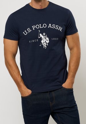 Man draagt een marineblauw U.S. Polo Assn. T-shirt met een wit polo spelerslogo en tekst, handen in de zakken van donkerblauwe spijkerbroek.