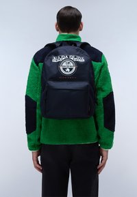 Mochila azul marino con un diseño redondeado, que presenta un bolsillo frontal y un logotipo. El modelo lleva una chaqueta de forro polar verde con detalles negros.
