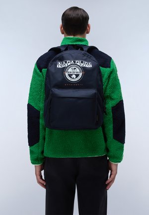 Zaino blu navy con un design arrotondato, dotato di una tasca frontale e un logo. Il modello indossa una giacca in pile verde con accenti neri.