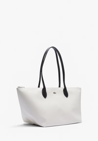 Tote bag in witte textuur met zwarte leren handvatten en accenten. Voorzien van een ritsluiting en een klein logo aan de voorkant.