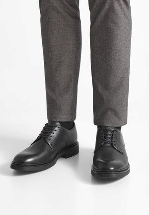 Schwarze Lederschuhe mit glatter Oberfläche, runder Zehenform und sechs Ösen. Getragen mit grauen karierten Hosen.