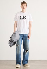 Witte katoenen T-shirt met vetgedrukt zwart "CK CALVIN KLEIN" logo, gecombineerd met lichtblauwe wijde versleten jeans en een lichtgrijze jas.