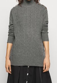 Pull à col roulé torsadé en gris chiné, avec manches longues et boutons décoratifs sur l'épaule. Tissu doux et texturé.