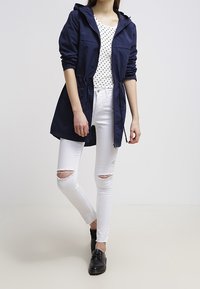 Veste imperméable marine avec capuche, taille à cordon, portée avec une chemise blanche à pois et un jean blanc déchiré, accompagnée de chaussures noires.