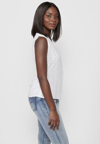 Haut sans manches en coton blanc avec des détails en œillets, présentant un devant boutonné et une coupe décontractée, associé à un jean en denim bleu.