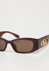 Ochelari de soare din plastic maro cu lentile de un maro închis. Prezintă un logo auriu "CK" pe braț. Design cu ramă rectangulară rotunjită.