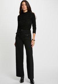 Donna che indossa un top a maniche lunghe con stampa leopardo nera, pantaloni neri a vita alta con tasche frontali, stivali neri alla caviglia, e bracciale e orecchini a cerchio dorati.
