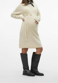 Personne enceinte portant une robe en tricot crème ceinturée, des bottes noires jusqu'aux genoux et un foulard noir noué autour du cou, se tenant les mains sur le ventre.