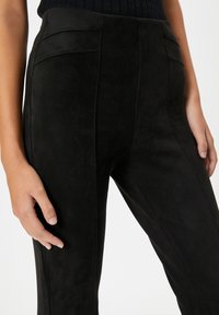 Pantalones de ante negro con una textura suave, que cuentan con dos bolsillos traseros y un ajuste elegante y entallado. Las manos descansan a los lados.