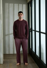 Langärmliges, dunkelrotes Hemd kombiniert mit maroon-weißen gestreiften Pyjamahosen. Einzelne Person steht auf Holzboden vor Glasfenstern im Hintergrund.