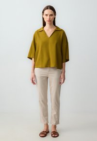 Olijfgroene oversized top met een V-hals en korte mouwen, gecombineerd met beige rechte broek en bruine slide-sandalen.