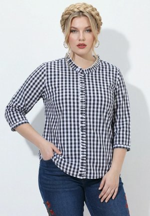 Kobieta z plecionymi blond włosami, ubrana w czarno-białą koszulę w kratę i niebieskie jeansy z czerwonym kwiatowym haftem, stojąca na białym tle.