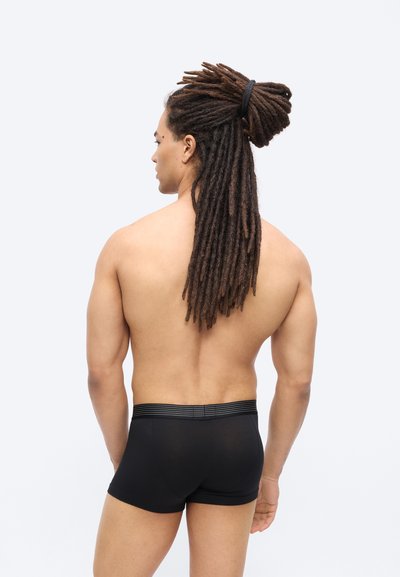 Άνδρας με μακριά πλεξούδες δεμένες πίσω, εμφανιζόμενος από πίσω, φοράει μαύρα εφαρμοστά boxer briefs σε απλό ανοιχτό φόντο.