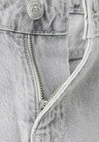 Gros plan sur un tissu denim gris avec une fermeture éclair métallique partiellement ouverte et un bouton marqué « Authentic Denim ».