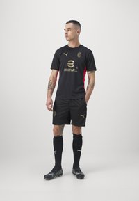 Svart fotbollskit med en gyllene logotyp och detaljer, med röda sidopaneler, tillsammans med svarta shorts och strumpor. Tillverkat av fukttransporterande tyg.
