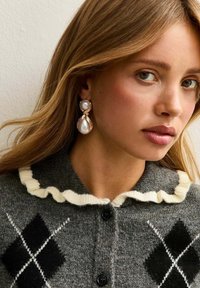 Guldnyans earrings med två texturerade pärlor, en rund och en droppformad, mot en bakgrund av en grå cardigan med en krämfärgad volangkrage.