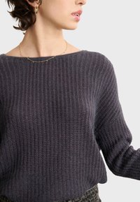Dunkellila, gerippter Strickpullover mit weitem Ausschnitt und langen Ärmeln, der über eine weiche Textur und eine lässige Passform verfügt. Akzent mit einer goldenen Halskette.