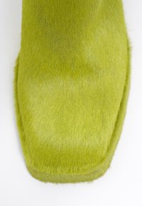 Punta de bota cubierta de piel de color verde con forma cuadrada, que presenta una textura suave y un borde liso, sobre un fondo blanco.
