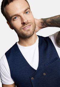 Granatowa kamizelka na białej koszulce, z zapięciem na dwa guziki i kieszonką na piersi, o gładkiej fakturze i dopasowanym kroju.