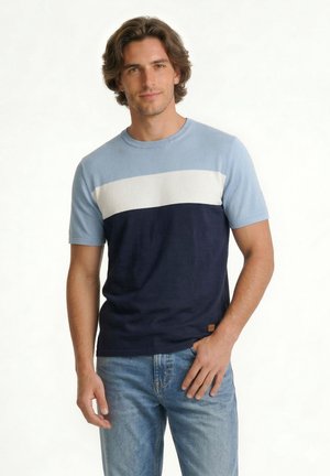 Bărbat care poartă un tricou cu mâneci scurte în blocuri de culori albastru deschis, alb și bleumarin, asortat cu blugi albastru deschis, stând cu o mână în buzunar.