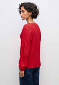 Blusa rossa, texture in rilievo con maniche lunghe a sbuffo. Presenta un collo arrotondato e un dettaglio con fiocco sul davanti. Abbinata a jeans blu scuro.