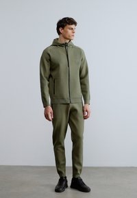 Sudadera con cremallera color caqui con paneles texturizados en los hombros, combinada con pantalones ajustados a juego. Se lleva con zapatillas negras; diseño minimalista.