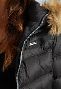 Zwarte pufferjack met een met bont gevoerde capuchon, voorzien van een zichtbare rits, gestikte panelen en een zwart label met de tekst "JACKIT".