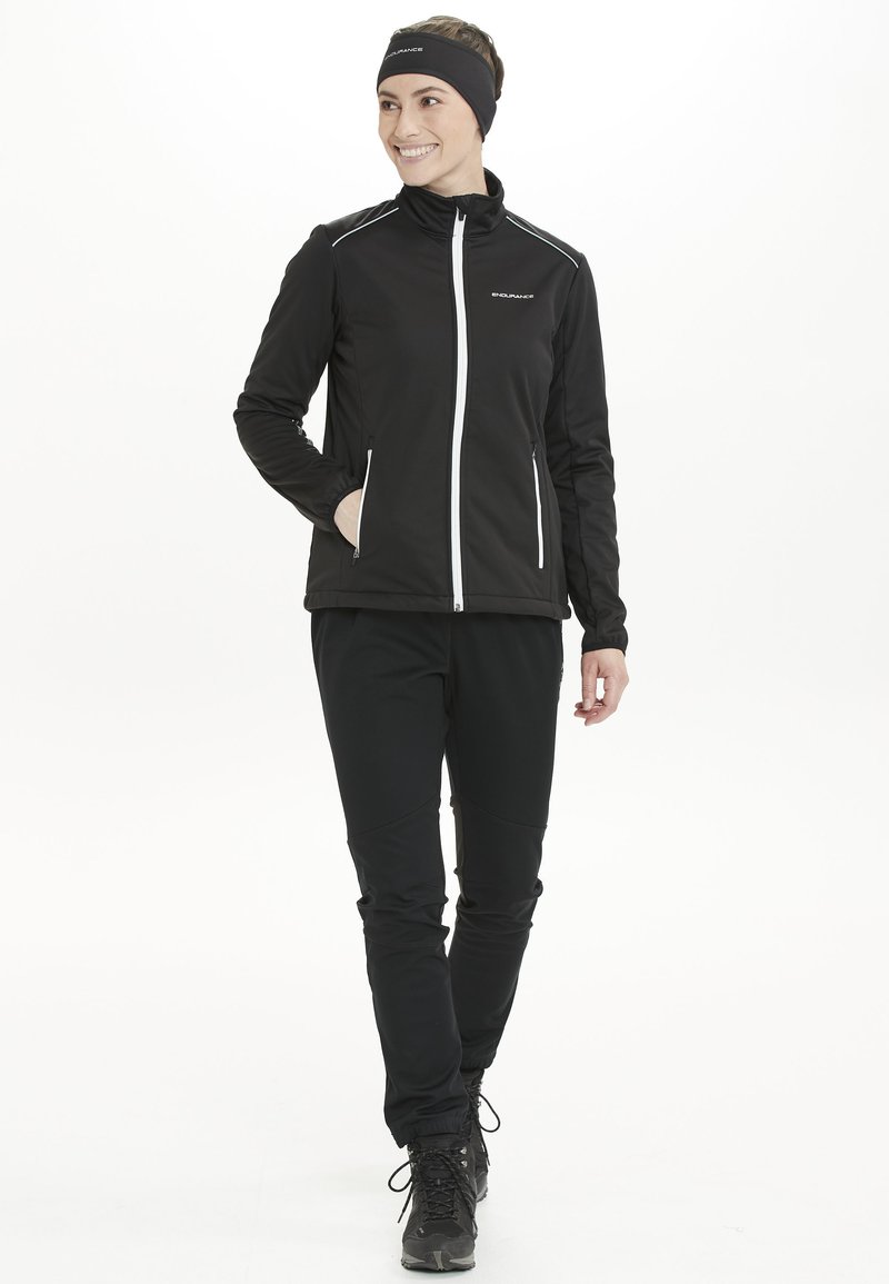 Sort atletisk jakke med hvide accenter og lynlåslukning, parret med sorte bukser. Glat tekstur, designet til udendørs aktiviteter, med lommer.