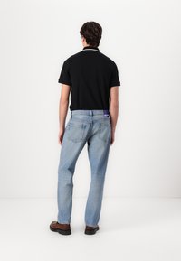 Schwarzes Polohemd mit kurzen Ärmeln, hellblaue gerade geschnittene Jeans, braune Schuhe und ein lila Etikett am hinteren Bund.