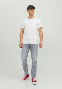 T-shirt en coton blanc avec une poche sur la poitrine, jeans gris clair et baskets rouges. Modèle portant une casquette noire ; fond uni.