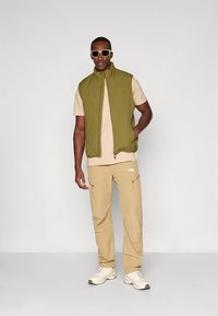 Mann trägt beige Sonnenbrille, olivgrüne ärmellose Weste, beige T-Shirt, beige Cargohose und weiße Sneakers, steht vor einfachem Hintergrund.