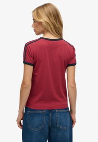 Bordeauxrotes T-Shirt mit marineblauen Einfassungen, kurzen Ärmeln und figurbetontem Schnitt. Die Rückseite zeigt minimale Details und einen abgerundeten Saum. Kombiniert mit blauen Jeans.