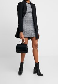 Manteau noir sur une robe grise à carreaux, bottines noires, et un petit sac à main noir avec une poignée transparente. Textures lisses et design ajusté.