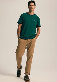 Zielony t-shirt z okrągłym dekoltem i granatowym wykończeniem, w połączeniu z beżowymi spodniami. Model ma na sobie ciemne buty z ozdobnym ściegiem.