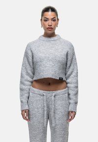 Vrouw met donker haar dat in een staart is gebonden, draagt een grijze, fuzzy cropped trui en bijpassende broek, staande tegen een eenvoudige witte achtergrond.