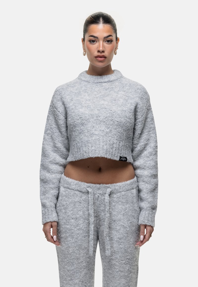 Vrouw met donker haar dat in een staart is gebonden, draagt een grijze, fuzzy cropped trui en bijpassende broek, staande tegen een eenvoudige witte achtergrond.