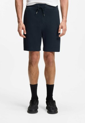 Man wearing navy drawstring shorts, black socks, and black sneakers, standing against a white background.